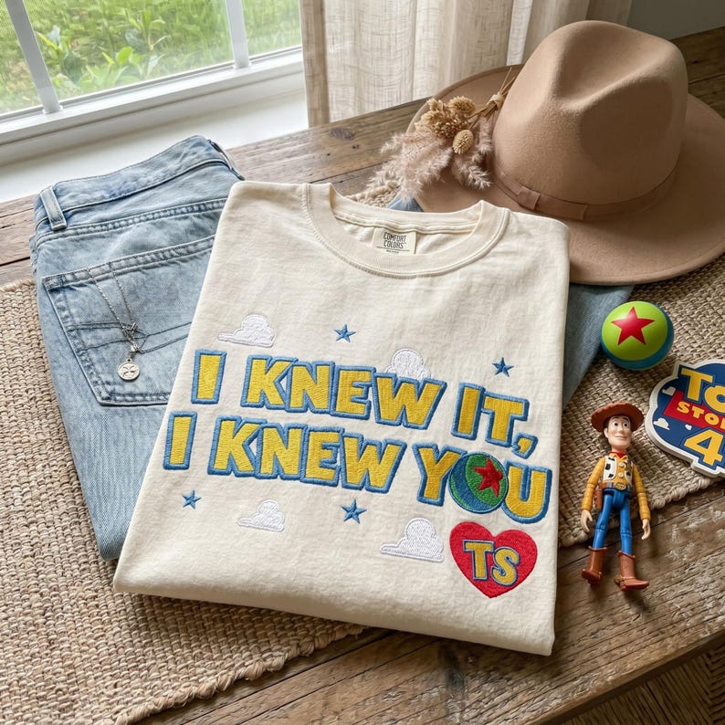 K&ouml;nnte beinhalten: Ein cremefarbenes T-Shirt mit gesticktem Text, der I KNEW IT, I KNEW YOU in Gelb mit blauen Umrandungen zeigt. Das Design enth&auml;lt kleine blaue Sterne, wei&szlig;e Wolken, einen bunten Ball mit einem roten Stern und ein rotes Herz mit den Buchstaben TS.