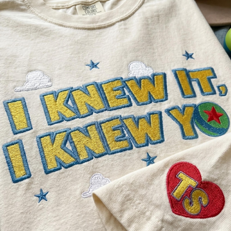 K&ouml;nnte beinhalten: Ein cremefarbenes T-Shirt mit gesticktem Text I KNEW IT, I KNEW YOU, erg&auml;nzt durch blaue Sterne, wei&szlig;e Wolken, einen gr&uuml;n-blauen Ball mit rotem Stern sowie ein rotes Herz auf dem &Auml;rmel, das die gelben Buchstaben TS enth&auml;lt.