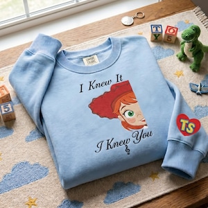 May include: A light blue crewneck sweatshirt featuring a graphic of a red-haired character with green eyes and the text "I Knew It I Knew You" with a treble clef symbol. The sleeve has a red heart patch with the yellow letters "TS" printed on it.