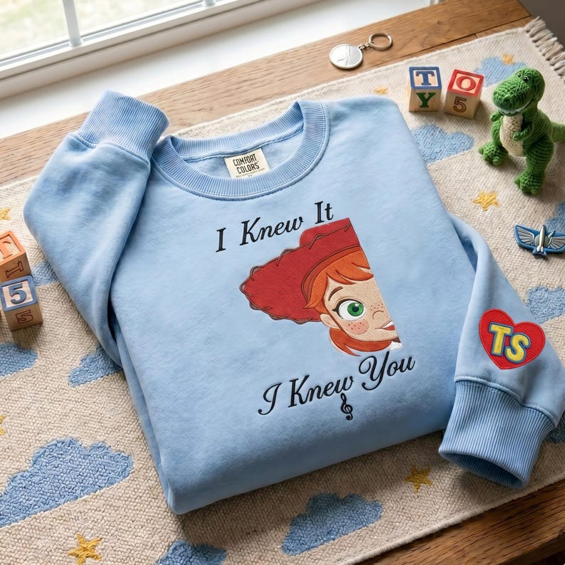 May include: A light blue crewneck sweatshirt featuring a graphic of a red-haired character with green eyes and the text "I Knew It I Knew You" with a treble clef symbol. The sleeve has a red heart patch with the yellow letters "TS" printed on it.