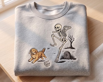 Puede incluir: Una sudadera gris claro de cuello redondo con un estampado gráfico de un perro golden retriever corriendo con un hueso en la boca, mientras un esqueleto baila cerca en un cementerio con una lápida y un árbol desnudo.