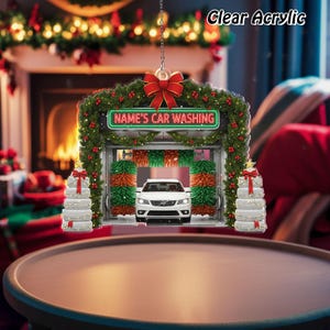 May include: Clear acrylic Christmas ornament featuring a car wash scene. The ornament includes a white car inside the car wash, a green and red wreath, and the text "NAME'S CAR WASHING". The ornament is decorated with a red bow and hanging chain.