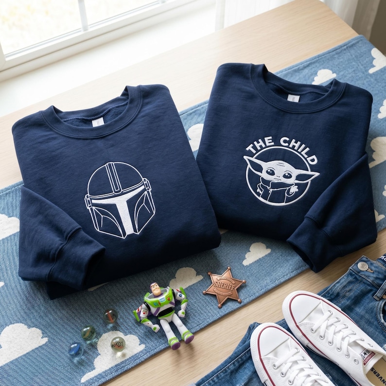 May include: Two navy blue crewneck sweatshirts. The left sweatshirt features a white line-art graphic of a Mandalorian helmet. The right sweatshirt displays a white circular graphic of Grogu with the text THE CHILD arched above the character.