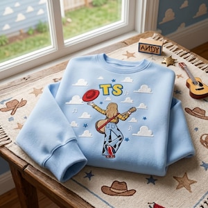 May include: A light blue crewneck sweatshirt featuring an embroidered graphic of a person with long hair playing a guitar, wearing a yellow plaid shirt and cow-print pants. The design includes a red hat, white clouds, blue stars, and the text TS.