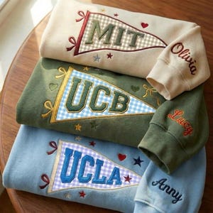 May include: A stack of sweatshirts in cream, green, and blue, each with a pennant design. The pennants display the letters "MIT", "UCB", and "UCLA" in different colors. Sleeves are embroidered with names. Stars and hearts decorate the sweatshirts.