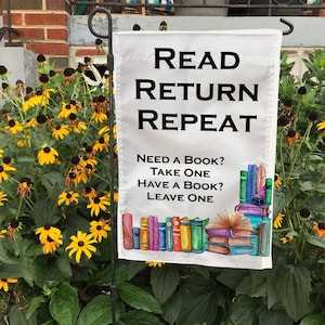 May include: White garden flag with the text "READ RETURN REPEAT" and "NEED A BOOK? TAKE ONE HAVE A BOOK? LEAVE ONE." The flag features a watercolor illustration of stacked books in various colors. The flag is displayed in a garden.