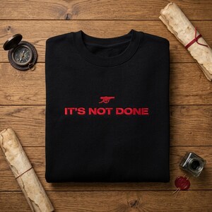 May include: Black crew neck sweatshirt with red embroidered text and logo. The text reads "IT'S NOT DONE" below a red cannon logo. The sweatshirt is displayed on a wooden surface with a compass, scroll, and inkwell.