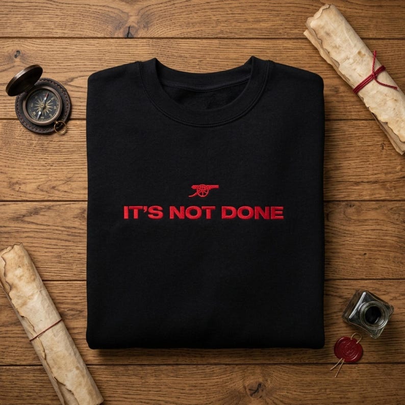 May include: Black crew neck sweatshirt with red embroidered text and logo. The text reads "IT'S NOT DONE" below a red cannon logo. The sweatshirt is displayed on a wooden surface with a compass, scroll, and inkwell.