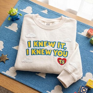 May include: A cream-colored crewneck sweatshirt featuring embroidered yellow text that reads "I KNEW IT, I KNEW YOU" with blue outlines. The design includes small white embroidered clouds and a red heart with the yellow letters "TS" inside.