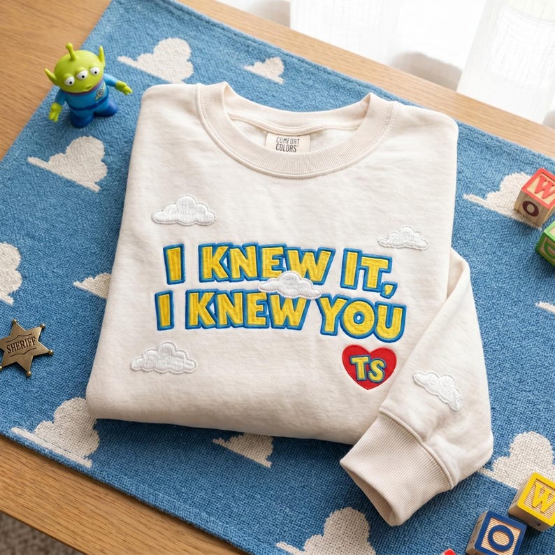 May include: A cream-colored crewneck sweatshirt featuring embroidered yellow text that reads "I KNEW IT, I KNEW YOU" with blue outlines. The design includes small white embroidered clouds and a red heart with the yellow letters "TS" inside.
