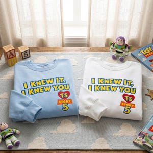 Puede incluir: Una sudadera azul claro y una sudadera blanca, cada una con el texto amarillo I KNEW IT, I KNEW YOU y un coraz&oacute;n rojo con las letras TS sobre el texto naranja STORY 5. Las prendas se muestran junto a art&iacute;culos tem&aacute;ticos de Toy Story.