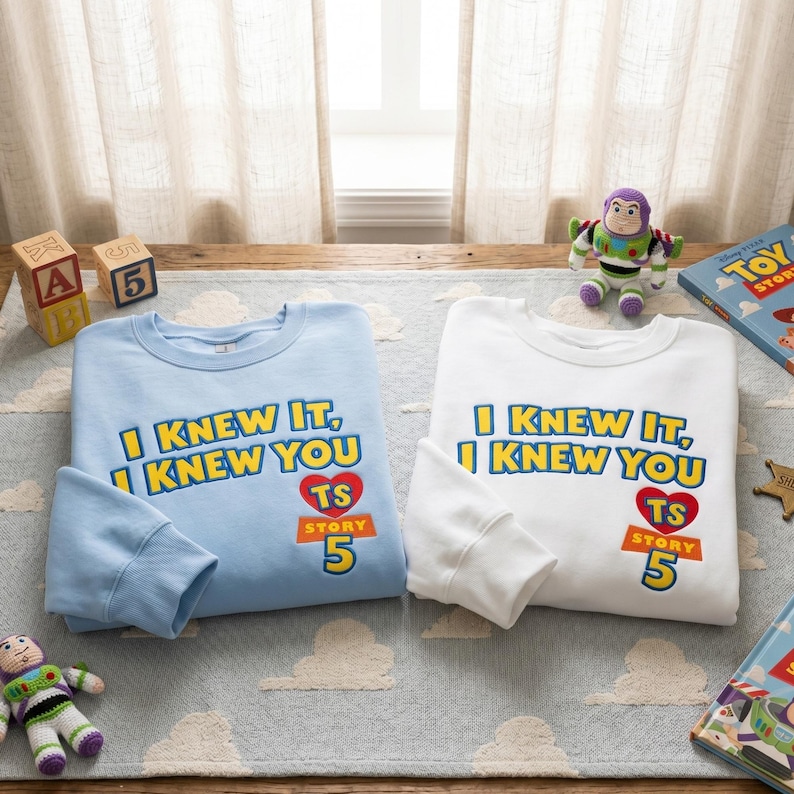 Puede incluir: Una sudadera azul claro y una sudadera blanca, cada una con el texto amarillo I KNEW IT, I KNEW YOU y un coraz&oacute;n rojo con las letras TS sobre el texto naranja STORY 5. Las prendas se muestran junto a art&iacute;culos tem&aacute;ticos de Toy Story.