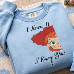May include: A light blue crewneck sweatshirt featuring a graphic of a red-haired character wearing a red cowboy hat. The text "I Knew It" is printed above the character, and "I Knew You" with a treble clef symbol is printed below the character.