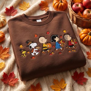 May include: A brown crewneck sweatshirt featuring an embroidered design of Snoopy, Woodstock, Charlie Brown, Lucy, and Linus dressed for autumn. The characters are depicted carrying pumpkins, a pie, and a basket of leaves amidst falling maple leaves.