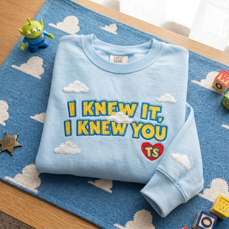 May include: A light blue crewneck sweatshirt featuring embroidered yellow text that reads I KNEW IT, I KNEW YOU, accompanied by a red heart with the letters TS inside. The design includes several white embroidered clouds on the front and sleeve.