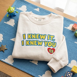 May include: A cream-colored crewneck sweatshirt featuring the embroidered text "I KNEW IT, I KNEW YOU" in yellow with blue outlines. Below the text is a red heart containing the yellow letters "TS". The garment has a ribbed collar and cuffs.
