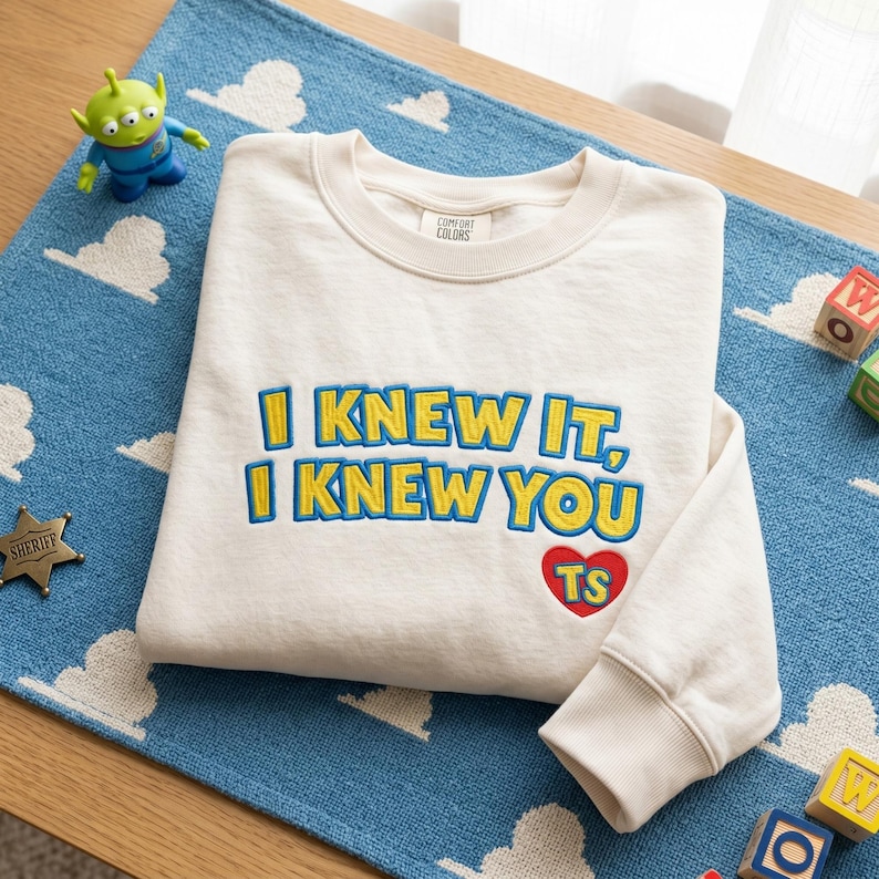 May include: A cream-colored crewneck sweatshirt featuring the embroidered text "I KNEW IT, I KNEW YOU" in yellow with blue outlines. Below the text is a red heart containing the yellow letters "TS". The garment has a ribbed collar and cuffs.