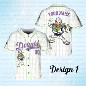 May include: White baseball jersey with green pinstripes and accents. Features the name "Donald" in purple script, the number "00", and a cartoon character. The back has "YOUR NAME" and another character. "Design 1" is written in black script.