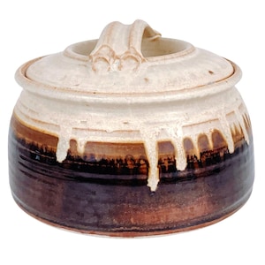 May include: A handcrafted ceramic jar with a lid. The jar features a two-tone design with a dark brown base and a cream-colored top. The lid has a handle. The glaze has a dripped effect, adding to the rustic aesthetic.