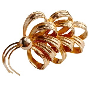 Może przedstawiać: Złota broszka w kształcie kokardy. Broszka posiada wiele pętli i centralną kulę, z delikatnymi zakrzywionymi akcentami. Metal ma teksturowany, żebrowany wzór, tworząc estetykę vintage.