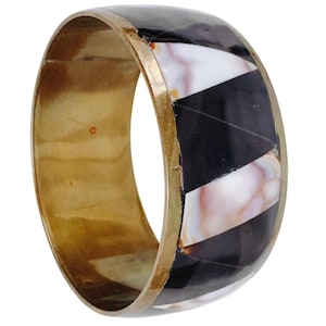May include: A wide, brass-toned bangle bracelet with a geometric design. The bracelet features alternating black, white, and pinkish-tan segments. The edges are trimmed with a thin, gold-colored band, creating a bold and stylish accessory.
