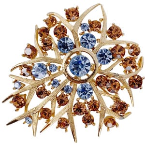 May include: Gold-toned brooch with a leaf-like design. The brooch features a central cluster of blue rhinestones surrounded by smaller blue and brown rhinestones. The brooch has a textured finish and is set against a white background.