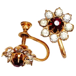 May include: A pair of vintage gold-toned screw-back earrings. Each earring features a central, deep red gemstone surrounded by clear, round crystals. The design resembles a flower. The earrings are set against a white background.