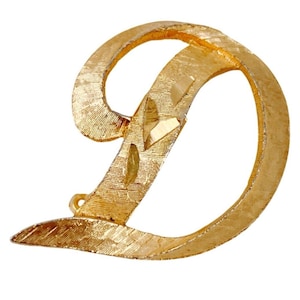 Broche con monograma en tono dorado, letra D, estilo manuscrito de mediados de siglo, firmado por Mamselle.