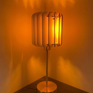 Może przedstawiać: Nowoczesna lampa stołowa z cylindrycznym kloszem wykonanym z pionowych drewnianych listew. Lampa ma podstawę i trzon w kolorze mosiądzu. Ciepłe światło żarówki tworzy miękką poświatę, tworząc przytulną atmosferę. Lampa jest podłączona.