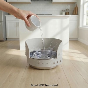 May include: A person pouring water into a light gray pet bowl with a stainless steel interior. The bowl has a decorative design and is sitting on a light wood floor. Water splashes as it hits the bowl.