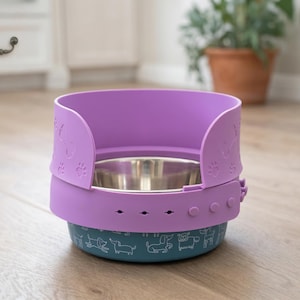 May include: A purple pet bowl with a stainless steel interior. The bowl has a teal base with white dog illustrations. The purple exterior features paw print cutouts and a protective shield. The bowl is designed to prevent spills.