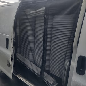 Puede incluir: Una furgoneta blanca con la puerta lateral abierta, que revela pilas de objetos cilíndricos grises. Hay una pantalla de malla negra. La furgoneta tiene una baca.