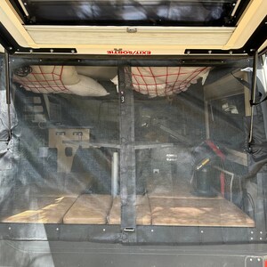 May include: A black mesh screen door with a red and white cargo net hanging above it. The door is open and the interior of the camper is visible. The text "EXIT/SORTIE" is printed on the top of the door.