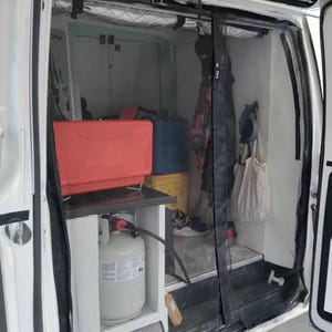 May include: A white van with a black mesh screen door. The door is open and the interior of the van is visible. There is a red petrol can, a white propane tank, and other items stored inside the van.