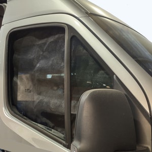 Puede incluir: Una sección del exterior de una furgoneta gris claro con una ventana lateral y una pantalla de malla negra. La ventana está enmarcada por un borde negro y el espejo lateral es visible. La imagen está tomada a la luz del día.