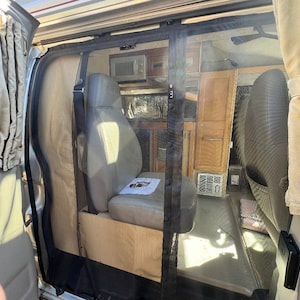 Op de afbeelding: Een zwarte gaasdeur is geïnstalleerd in een bestelwagen. De deur staat open en het interieur van de bestelwagen is zichtbaar. Het interieur is afgewerkt met houten panelen en heeft een magnetron, kasten en een stoel.