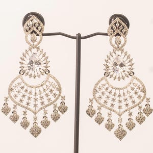 May include: A pair of silver-toned chandelier earrings. Each earring features a teardrop-shaped clear gemstone at the center, surrounded by smaller clear stones. The earrings have a detailed, openwork design with dangling heart-shaped accents.