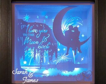Könnte beinhalten: Ein quadratischer Schattenkasten mit schwarzem Rahmen zeigt eine blau hinterleuchtete Szene eines Paares auf einer Mondsichel. Der Text lautet I love you TO THE Moon & back, Sarah & James und Personalization Avaliable. Silhouetten einer Weide, Feuerwerk und zwei Herzen sind zu sehen.
