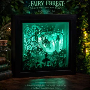 May include: A black square shadow box featuring a layered paper cutout scene of a forest with fairies, mushrooms, and trees. The interior is illuminated with a bright green light. Text at the top reads FAIRY FOREST SHADOW BOX.