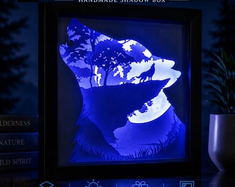 May include: A square black shadow box frame containing multi-layer paper art illuminated by soft blue LED light. The artwork depicts a wolf howling on a cliffside against a large moon and forest silhouette. Text reads WOLF IN THE WILD.