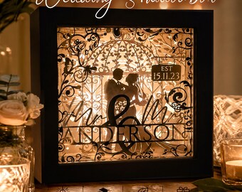 Könnte beinhalten: Ein schwarzer, quadratischer Schattenkasten mit einem mehrschichtigen Papierschnitt-Design eines Paares in einem Pavillon. Die Silhouette enthält den Text Mr & Mrs ANDERSON und EST 15.11.23. Das Display ist mit warmem LED-Licht beleuchtet.