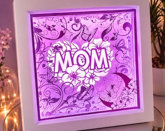 Könnte beinhalten: Ein quadratischer weißer Schattenfugenrahmen mit einem mehrschichtigen Papierkunstwerk, das in violettem Licht erstrahlt. Das Design zeigt das Wort MOM in der Mitte, umgeben von komplizierten Blumenmustern, Schmetterlingen und einer Mondsichel.