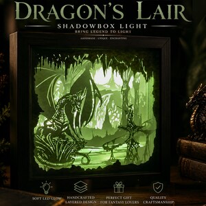 May include: A square black shadowbox light featuring a layered paper cutout scene of a dragon and a figure in a cavernous landscape. The interior is illuminated with a soft green LED glow. Text reads DRAGON'S LAIR SHADOWBOX LIGHT BRING LEGEND TO LIGHT.