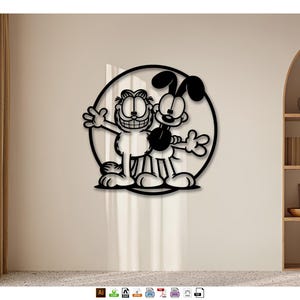 Peut inclure: Décoration murale en métal noir représentant les personnages de dessins animés Garfield et Odie à l'intérieur d'un cercle. Les personnages sourient et saluent. L'œuvre est fixée sur un mur beige.