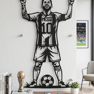 Puede incluir: Una silueta de arte de pared de metal negro que representa a un jugador de fútbol con los brazos levantados en celebración, vistiendo una camiseta con el nombre MESSI y el número 10. La figura se encuentra sobre un balón de fútbol con detalles.