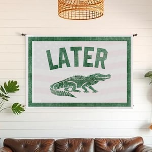 May include: A brown leather sofa with three cushions sits below a wall hanging. The wall hanging has the word "LATER" in green letters above a green alligator illustration. The background is white.