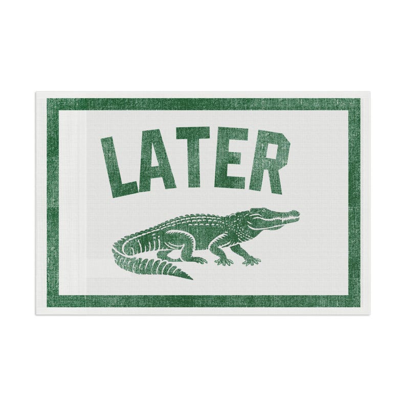 May include: A rectangular, white sign with a green border and text. The word "LATER" is printed in large, green, uppercase letters above a green alligator illustration. The design has a distressed, vintage look.