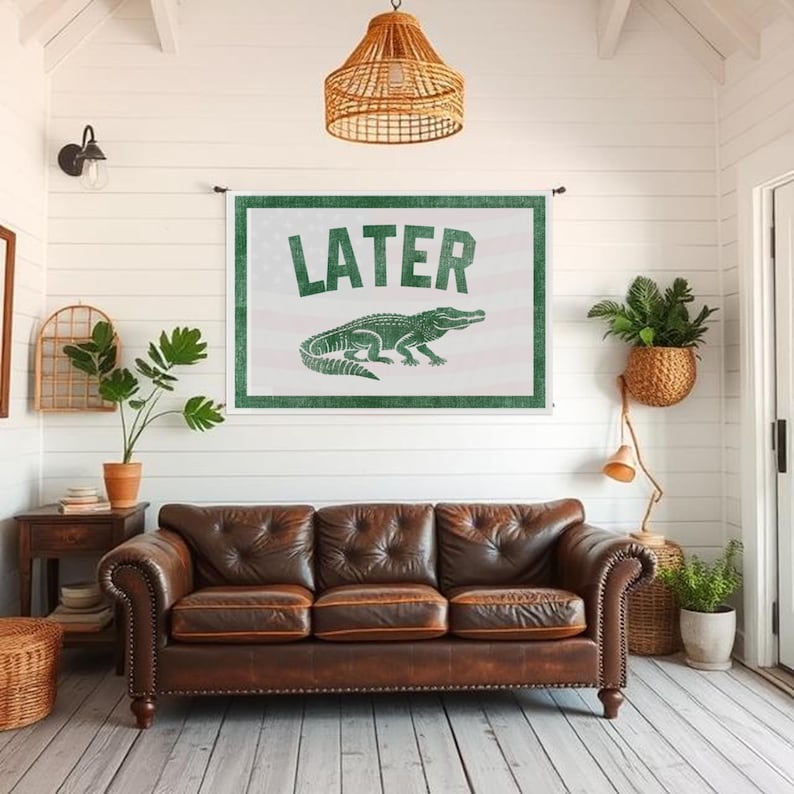May include: A brown leather sofa with three cushions sits below a wall hanging. The wall hanging has the word "LATER" in green letters above a green alligator illustration. The background is white.