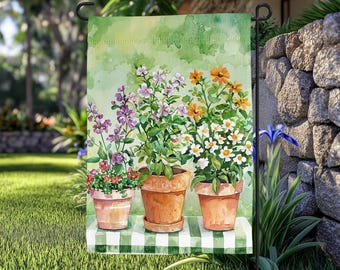 May include: A garden flag depicting a watercolour painting of three terracotta pots filled with colourful flowers. The flowers include purple, orange, and white blooms. The flag is set against a green background and a green and white checked table.