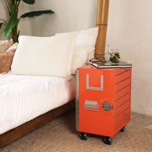 May include: An orange bedside table with a metal handle and accents, resting on small black wheels. The table has a rectangular shape with a series of horizontal lines. A book, glasses, and a glass of tea sit on top.